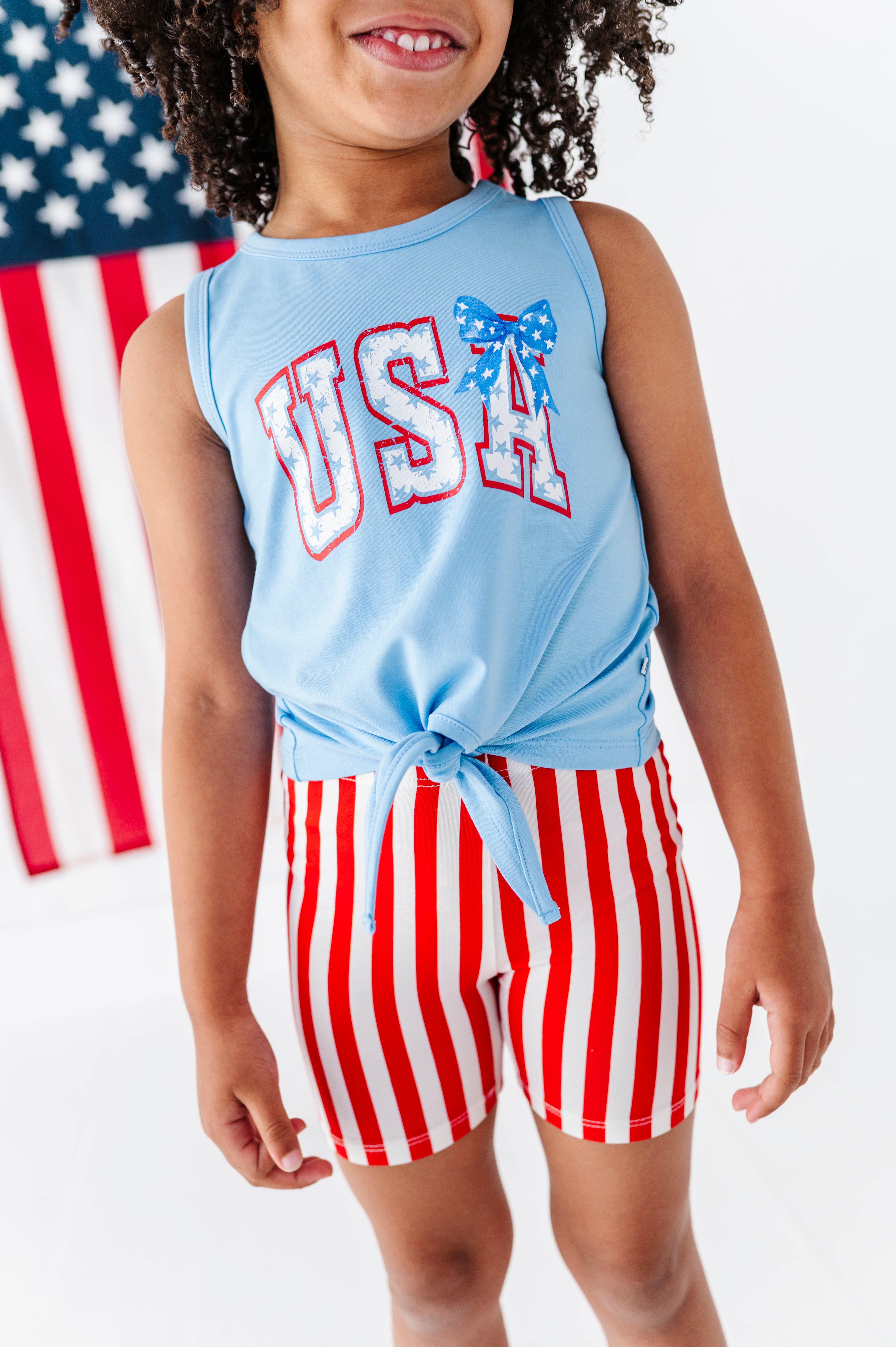 Child wearing a light blue sleeveless tank top with 'USA' and red and white striped bamboo biker shorts in front of an American flag.